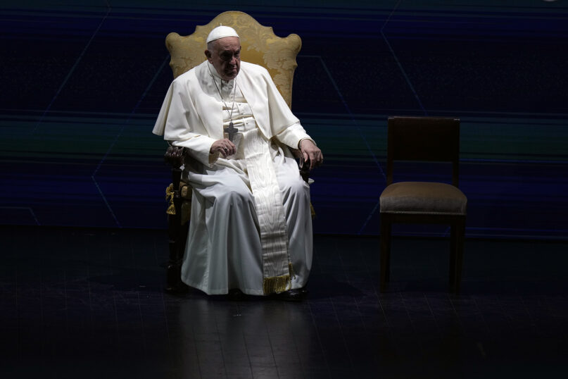 Francis sits on chair, highlighted by light, surrounded by shadow.