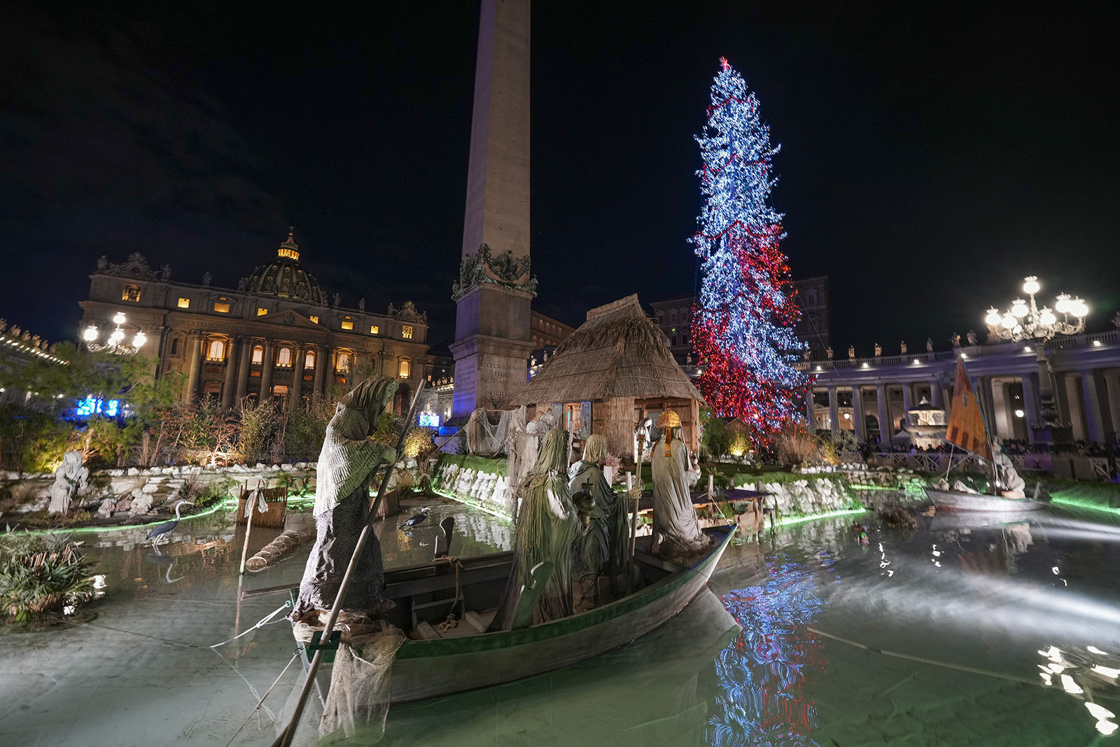 Illuminated tree and Vatican obelisk visible in background; foregrounded is part of nativity scene. 