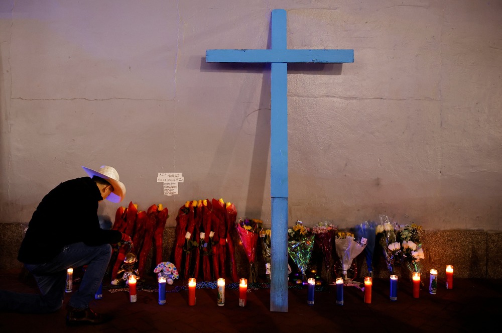 A cross leans against a wall lined with votive candles, on left a man kneels.