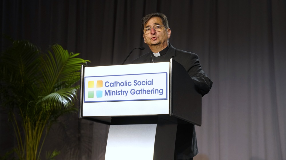 The bishops wears plain black clerics and stands speaking at podium.