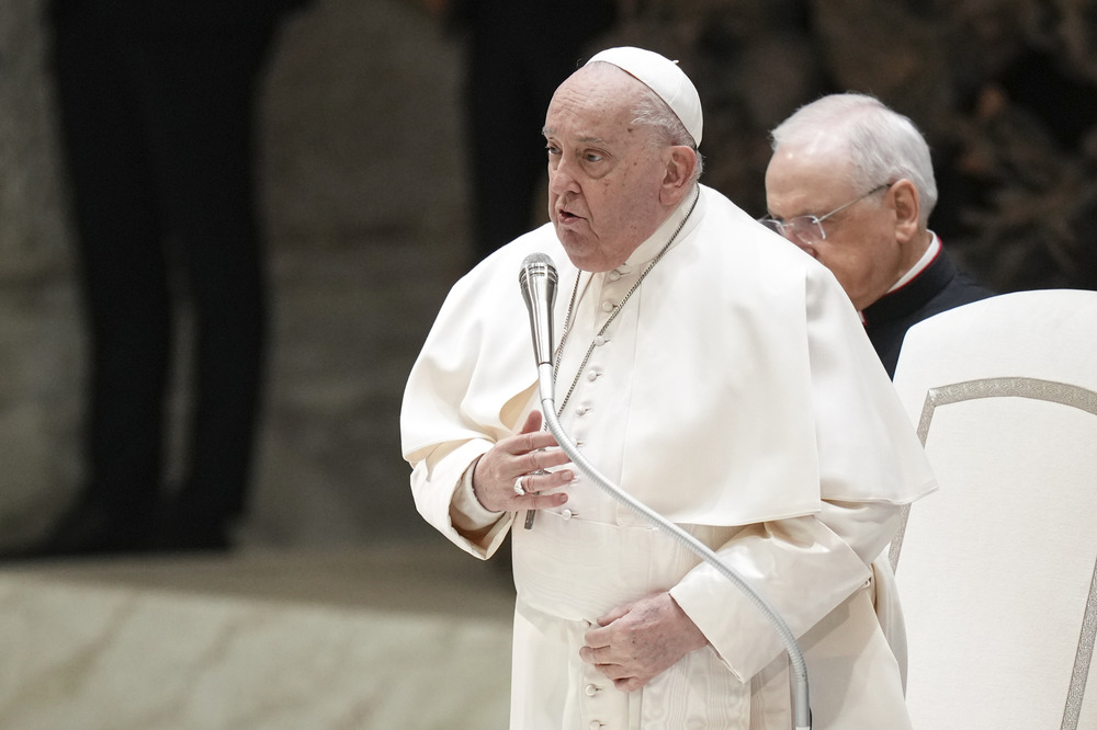 The pope stands speaking into microphone with hand to chest.