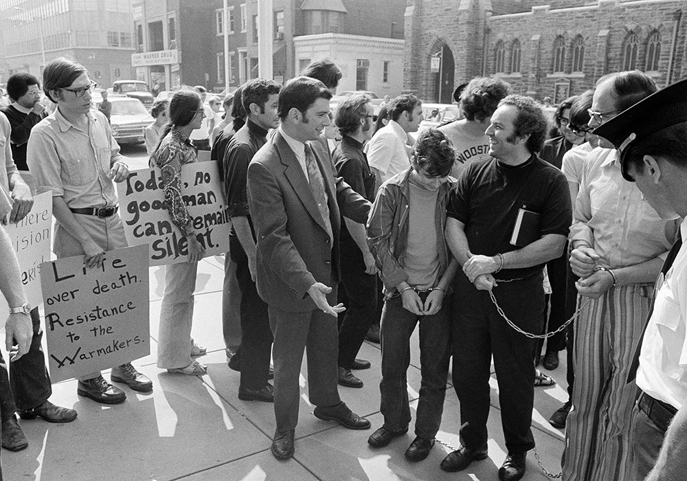 Supporters of persons arrested a week earlier in an attempted raid on a Selective Service office greet those arrested at a court hearing in Camden, New Jersey, on Aug. 30, 1971. Those arrested were brought from a New Jersey jail in handcuffs. Among them is John Grady, right, handcuffed, wearing black.  They became known as the "Camden 28." (AP photo/Bill Ingraham)