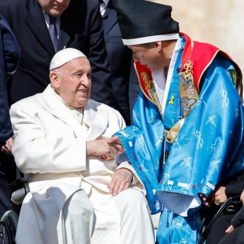 Pope Francis, from wheelchair, shakes hands with audience wearing vibrant, traditional garb.