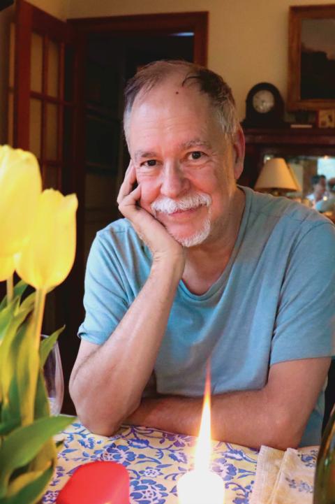 Gregory Macguire, author of Wicked: The Life and Times of the Wicked Witch of the West, is pictured from the chest-up, seated and subtly smiling with his arms on a table.