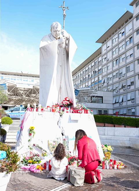 A woman and a young girl pray at the base of a statue of St. John Paul II outside Rome’s Gemelli hospital on March 2. (CNS/Lola Gomez)