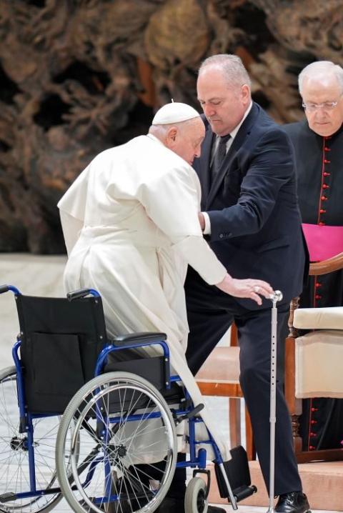 Pope Francis, assisted by an aide, rises from his wheelchair to take his seat in the Paul VI Audience Hall.