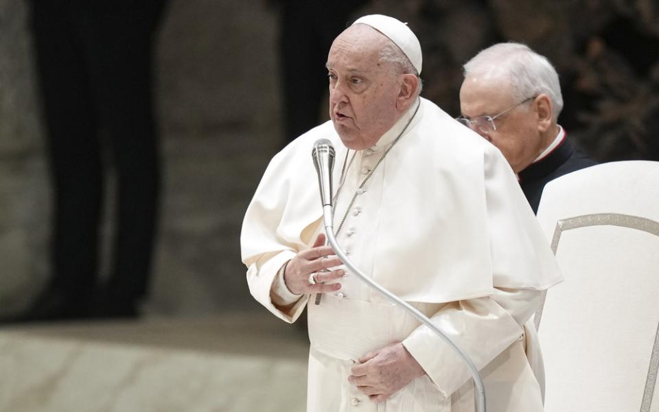 The pope stands speaking into microphone with hand to chest.