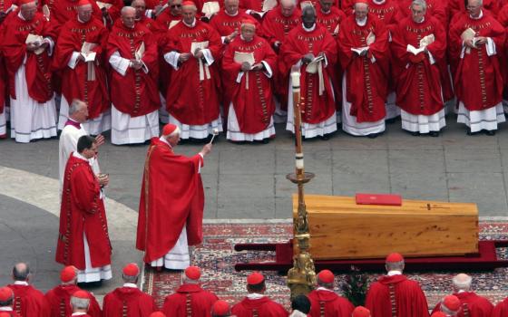 Many cardinals wearing red surround a wooden casket on a rug in St. Peter's Square