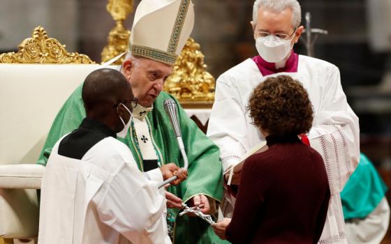 Pope Francis, wearing a mitre and no mask, is flanked by two masked priests and hands a crucifix to a masked woman