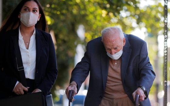 McCarick wears a suit and suit and hunches over a walker. A young woman wearing a mask stands next to him.