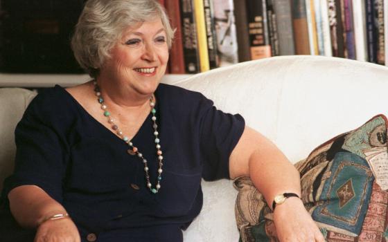 An older white woman wearing a blue dress and a beaded necklace sits on a white couch in front of a bookshelf