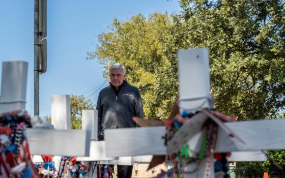 An older man in a quarter zip sweater stands before white crosses outside