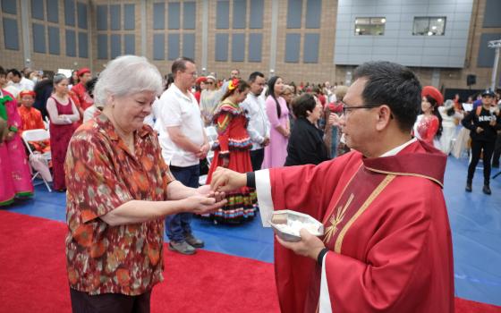 A black-haired man wearing glasses and a red chasuble places a Communion wafer in an older woman's hands