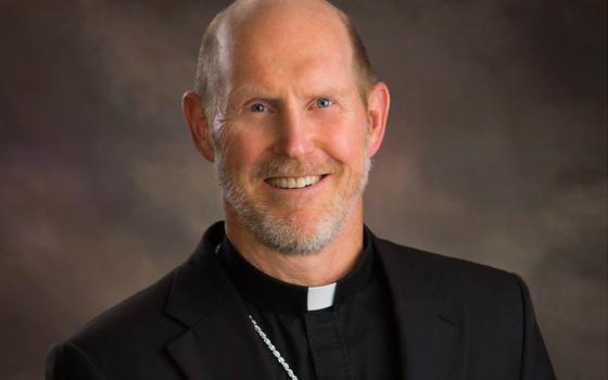 A white man with blue eyes and a beard wears a clerical collar and smiles at the camera