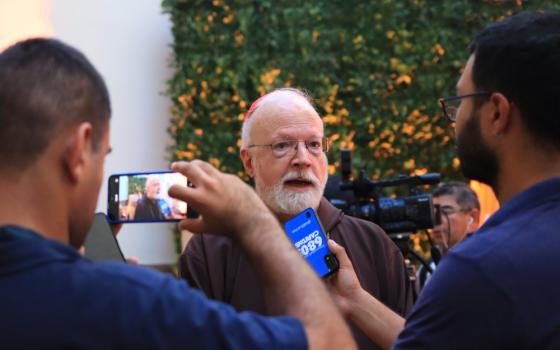 Two people hold phones towards an older white man wearing a brown robe and a red zucchetto