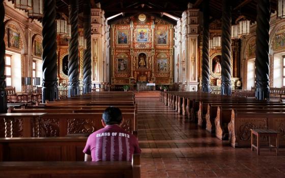 Man pictured from behind, seated in rear pew; aisle, columned nave, and elaborate altar of cathedral visible. 