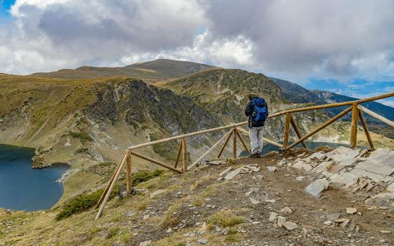 A person stands before a wooden barrier in a landscape with mountains and a lake (Unsplash/Luba Ertel)