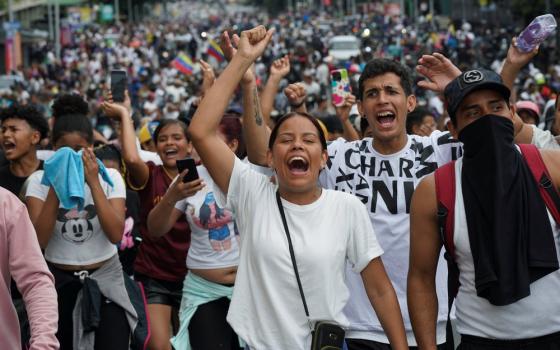 Woman in white shirt shouts chant while raising fist; behind hundreds of white-clothed people assemble and march. 