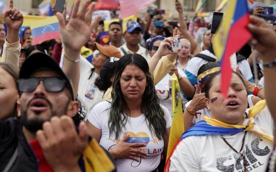 Woman in center of frame, surrounded by large crowd; Venezuelan flag colors on garments, demonstration items and flags. 