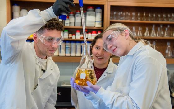 Three students wear white lab coats, and observe as one student uses a dropper to insert fluid into Erlenmeyer flask. 