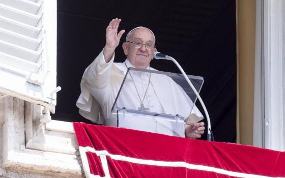 Pope raises hand in greeting, standing at lectern in window of Apostolic Palace. 