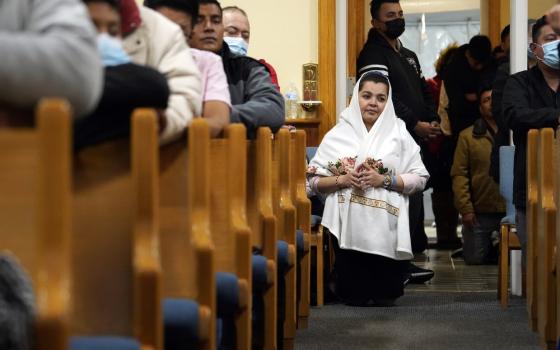 Woman wears white mantle and kneels in church aisle; others kneel in pews. 