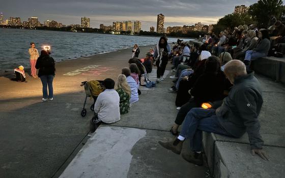 Chicago skyline at dusk in background; people sit gathered around speakers at edge of Lake Michigan. 