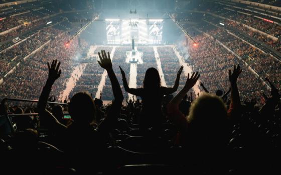 Back view of people in nosebleed seats raising hands; light emanates from distant stage. 