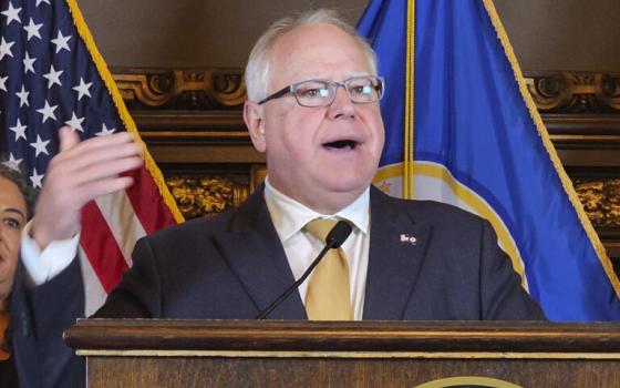 Gov. Walz stands at podium speaking, behind are US and Minnesota flags. 