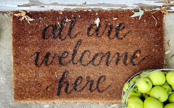 A welcome mat that says "All are welcome here" sits next to a basket of green apples (Unsplash/Leeann Cline)