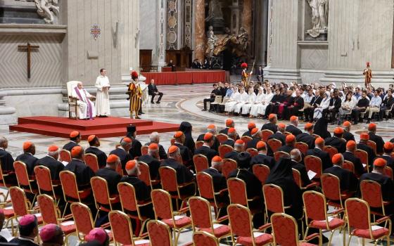 Pope seated on red dais; to his left is a group of young people, and to his right facing straight ahead are rows of prelates.