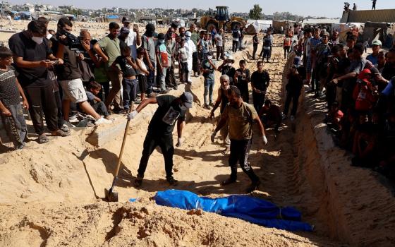 People line up along a ditch, standing in the ditch are the diggers, and wrapped in a blue tarp is a corpse for burial.