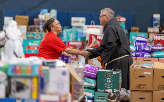 The bishop, wearing habit, shakes hands with a woman. The two stand amid large stockpile of supplies.