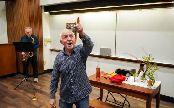 Sandford looks up and out pointing, behind him is classroom whiteboard and Benoit with saxophone.