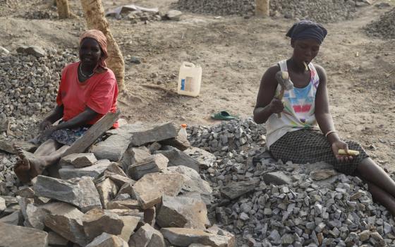 Two women sit on a pile of rocks holding mallets.