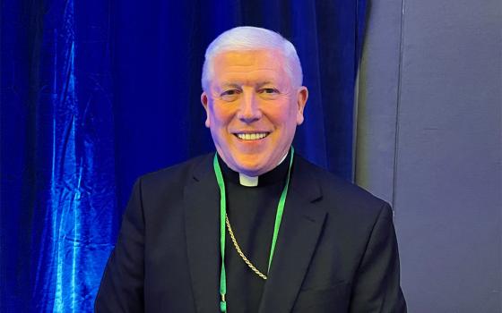 The bishop wears plain black clerics and smiles against a blue backdrop.