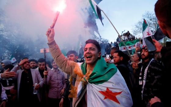 Overjoyed young man wearing Syrian flag, holds flare and smiles; behind him is crowd.