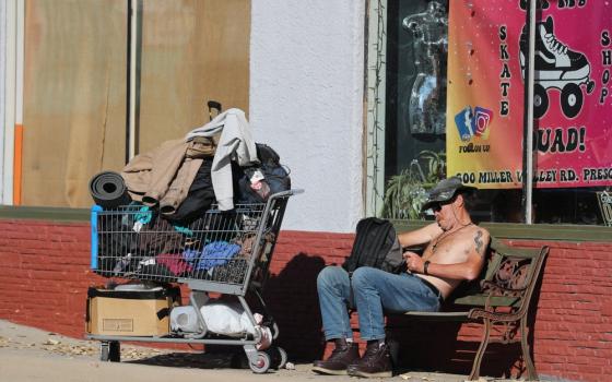 Man sits shirtless on bench, next to him is shopping cart filled with posessions.