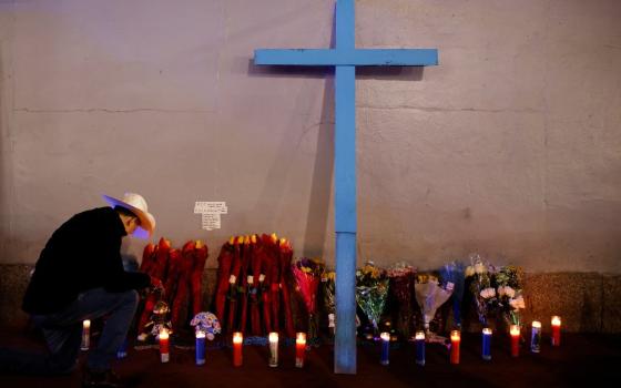 A cross leans against a wall lined with votive candles, on left a man kneels.