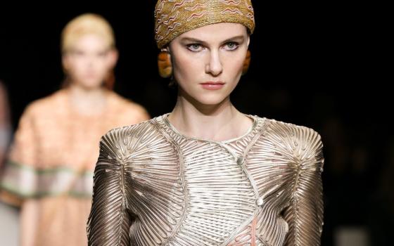 Model on runway foregrounded staring off camera, model behind seen out of focus. Garments metallic and earth-toned.