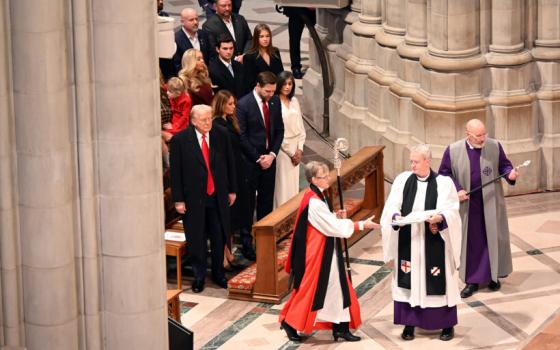 Bishop Budde wearing choir dress and holding large crozier processes past Trump.