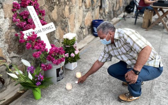 A man in a mask kneeling to light a candle before a wooden cross in a bucket of pink flowers and bearing the name of his sister