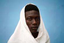 Young man, draped in white raiment, peers poignantly into camera; behind him is blue background.