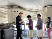 Beneath a tent held up by water tanks, the scholastic stands in front of a line of people.